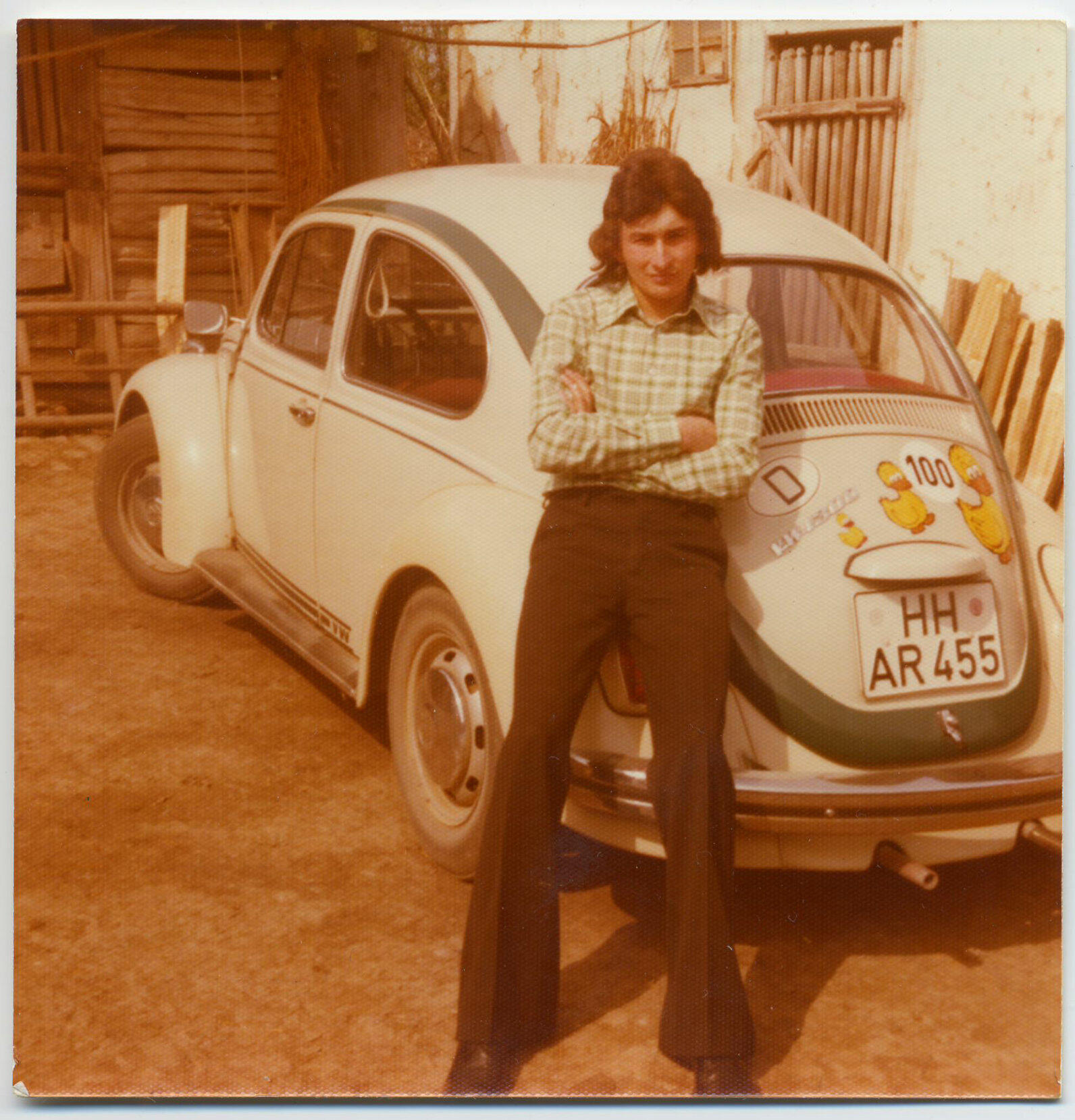 Ein Farbfoto zeigt einen jungen Mann mit verschränkten Armen, der vor einem weißen VW Käfer lehnt. Der Mann trägt ein kariertes Hemd und dunkle Hosen. Der Käfer hat Hamburger Kennzeichen und Aufkleber auf der Rückseite.