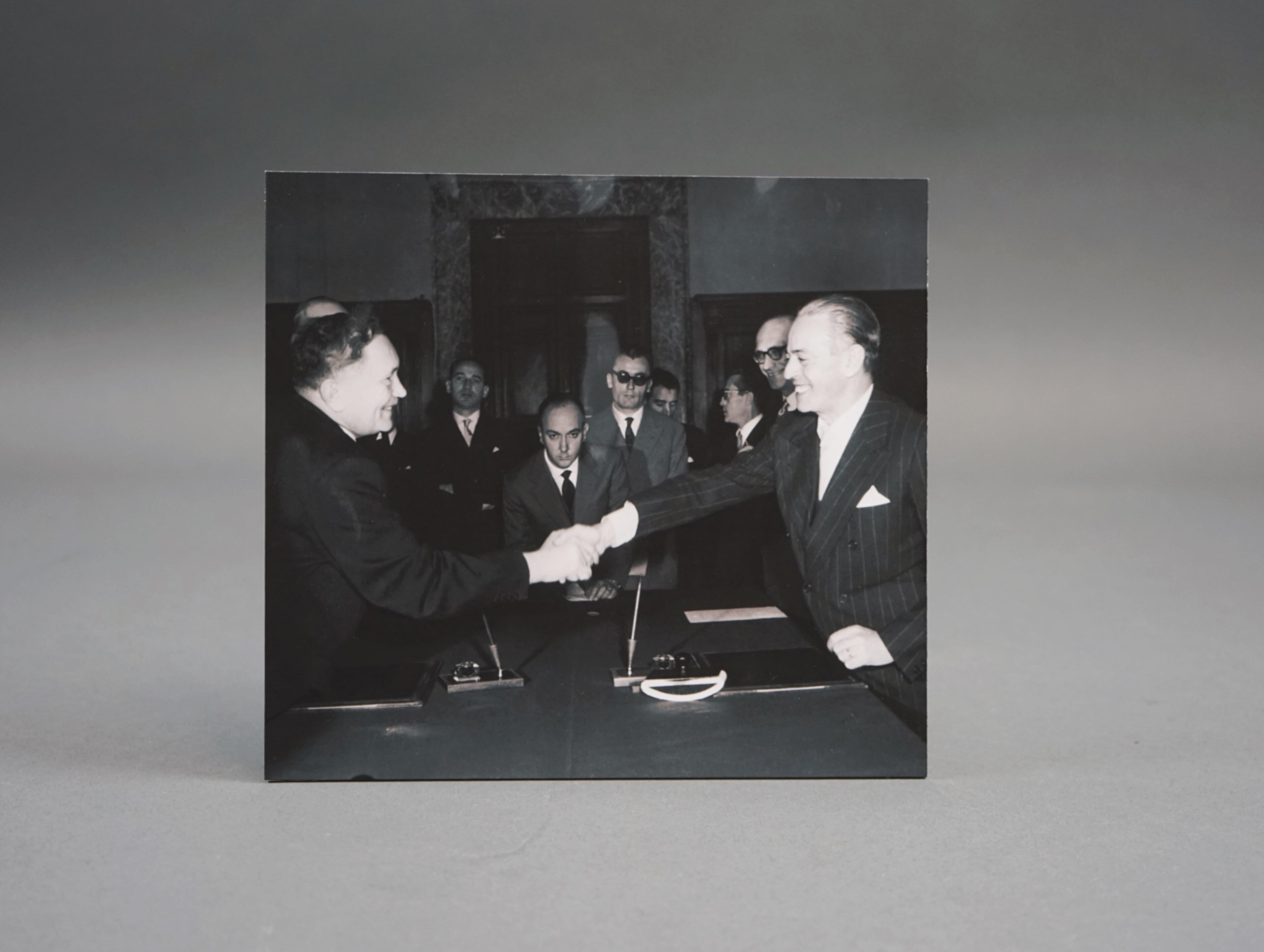 A black and white photograph of two men in suits shaking hands across a table. In the background are other men in suits. The atmosphere of an official occasion.