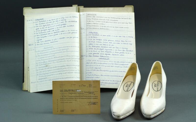 An open book with handwritten notes, a report book, in front of it a form on dark cardboard and a pair of white wedding shoes.