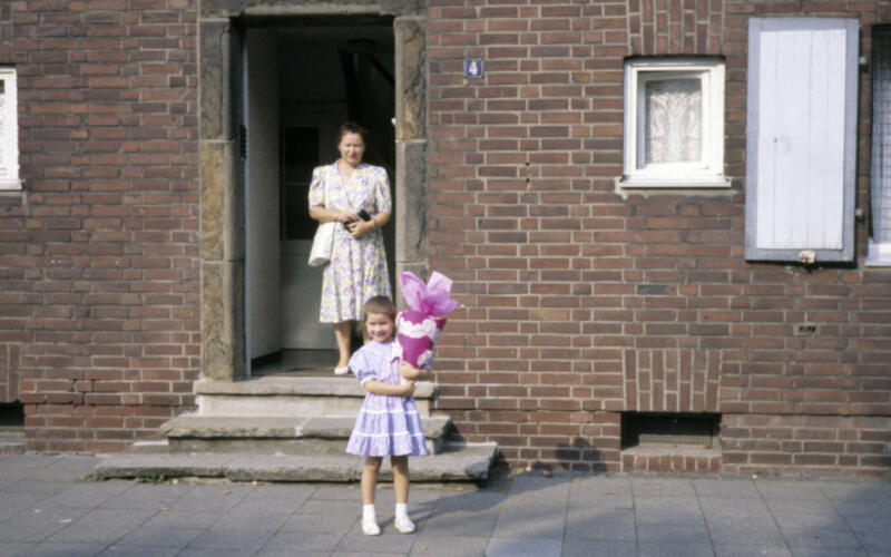 Ein Farbfoto zeigt ein kleines Mädchen in einem lila Kleid, das auf dem Bürgersteig vor einem Backsteinhaus steht und eine Schultüte hält. Hinter ihr, in der offenen Haustür, steht seine Mutter in einem geblümten Kleid.