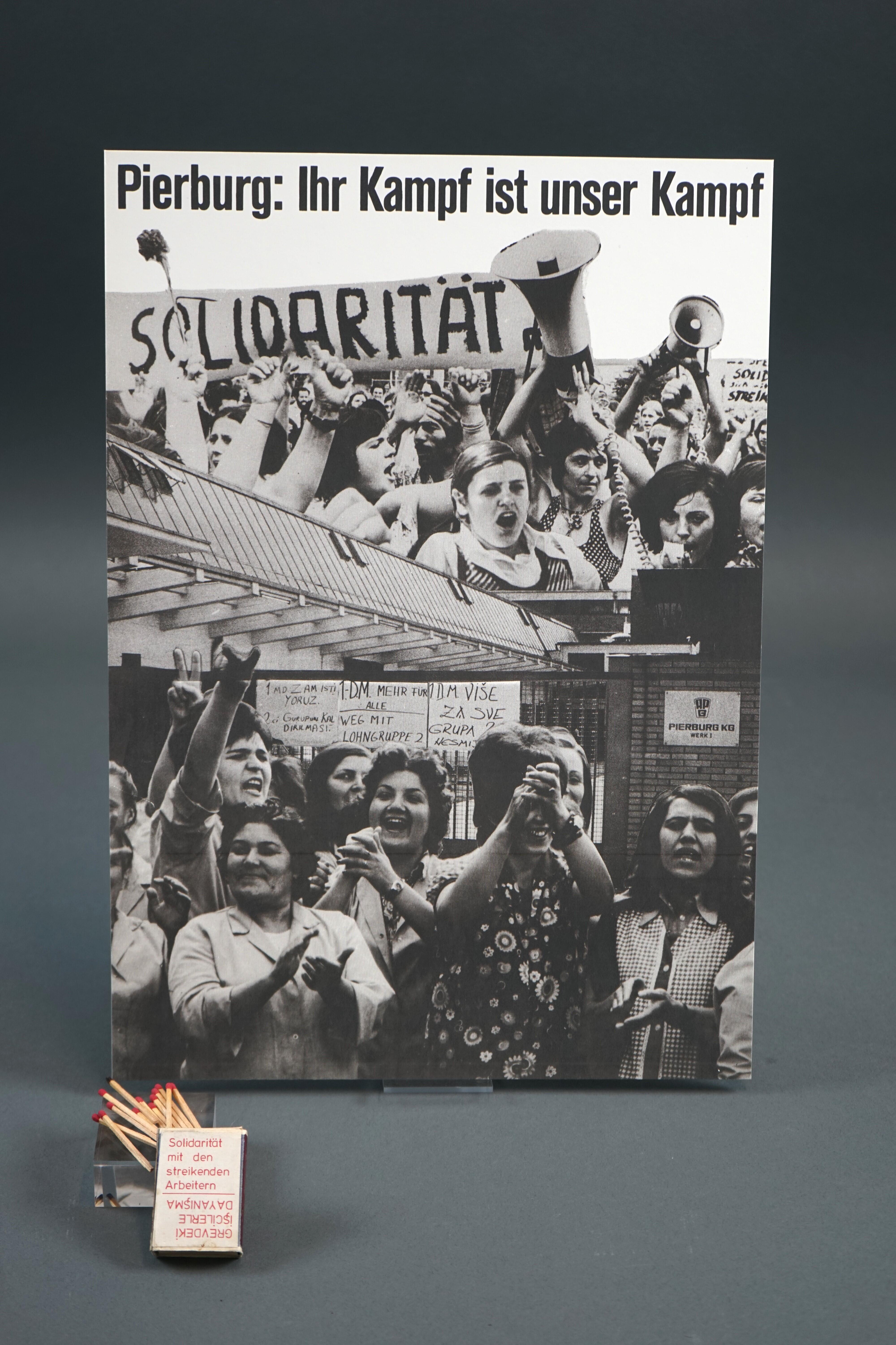 A film poster from 1974 with the inscription ‘Pierburg: Your fight is our fight’. A matchbox from the early 1980s with the inscription ‘Solidarity with the striking workers’ is placed in front of it.