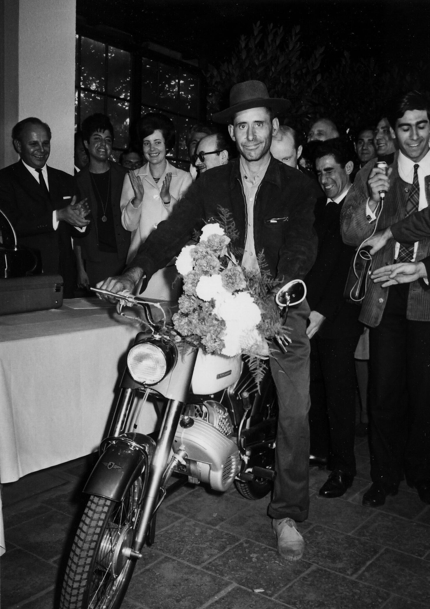 A black and white photograph of a man sitting on a moped, smiling slightly uncertainly at the camera. Other people are standing around him.