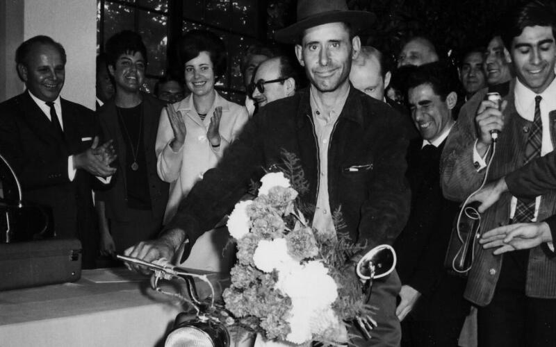 A black and white photograph of a man sitting on a moped, smiling slightly uncertainly at the camera. Other people are standing around him.